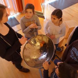 Die Gruppe steht um eine Steel Pan. Die Pädagogin erklärt das Instrument. Die kinder schauen interessiert zu ihr auf.