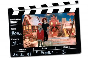 Auf einer Filmklappe ist ein Foto mit mit einem Wunderfinder-Kind und einer Parin, die mit roten Mantel und Hüten verkleidet in einer Winterlandschaft stehen.